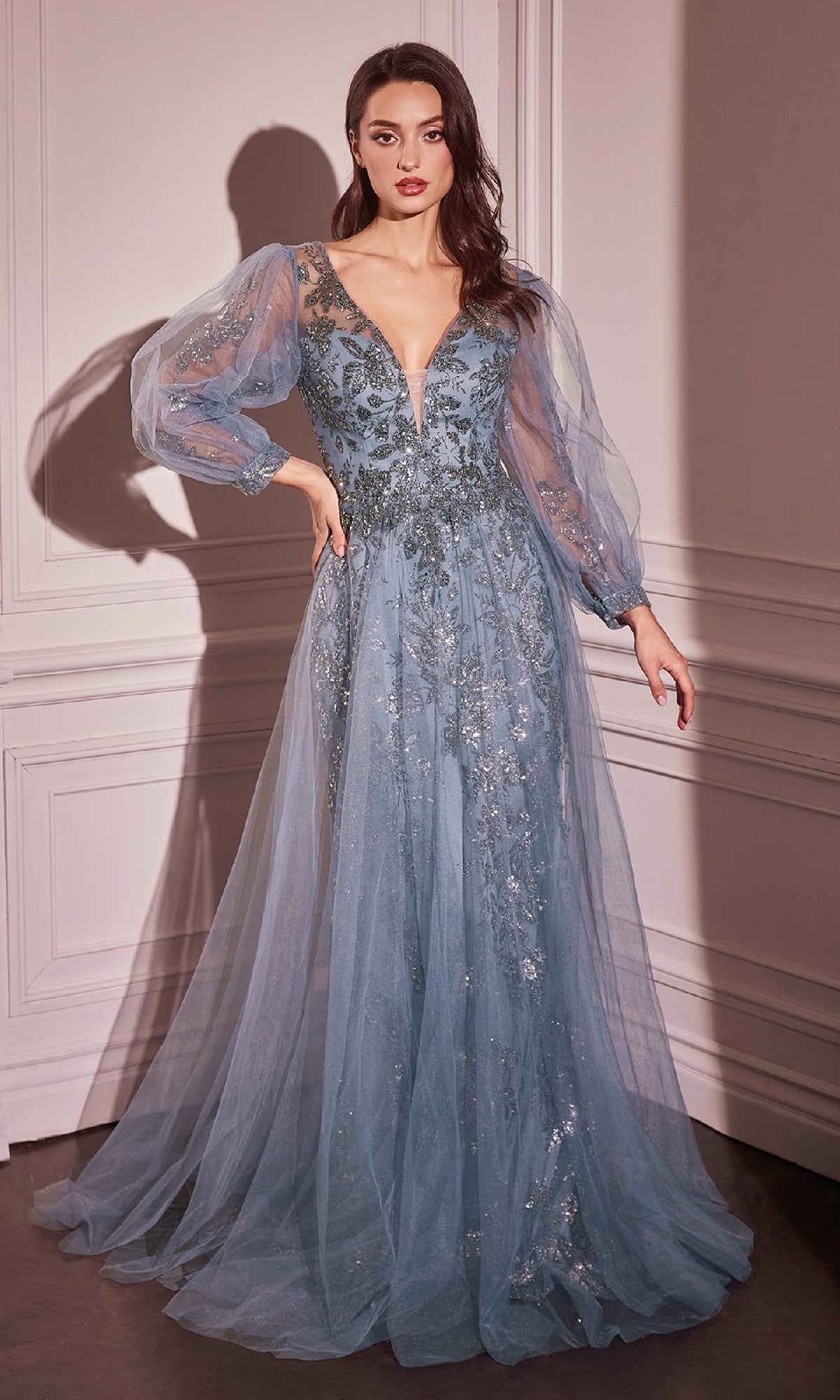 A woman stands in an elegant room wearing the Ladivine CD0102 Sheer-Sleeve Long Prom Dress, a floor-length blue gown featuring floral embellishments, long puffed sleeves, and a deep V-neckline.